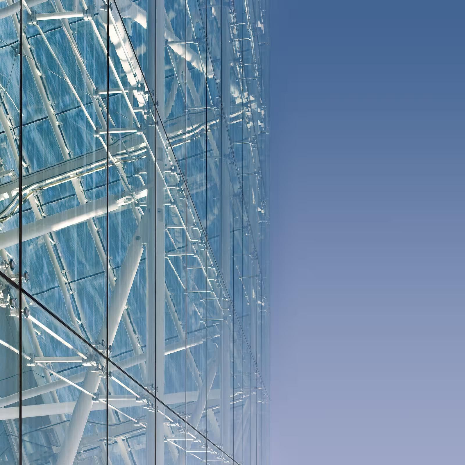 The glass facade of a building, with the structural framework visible behind the glass, is seen against a blue sky.